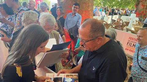 Die Schauspielerin Meret Becker steht beim Festival des deutschen Films in Ludwigshafen auf dem Roten Teppich und schreibt einem Mann ein Autogramm in eine Zeitschrift.