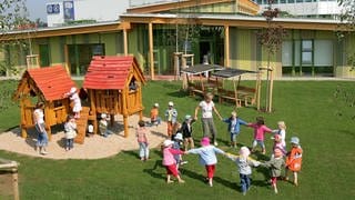 Kindertagesstättengebäude mit Kindern und Erzieherin - Symbolbild für Kreise müssen mehr für Kindergarten-Gebäude bezahlen