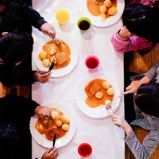 Kinder essen an Tisch mit bunten Bechern (Symbolbild). Die Aktion Kindervesperkirche wird dieses Jahr in Ludwigshafen auf zwei Wochen verlängert.