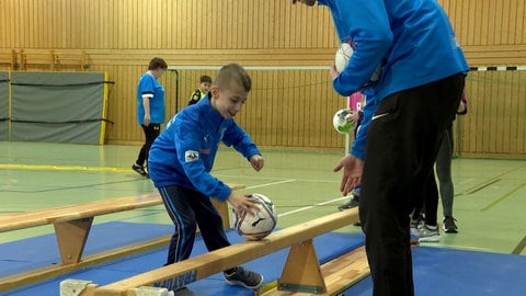 Ein Kind im blauen Trikot der TSG Deidesheim schiebt einen Ball über eine umgedrehte Bank. Die Kinder sind sehr kreativ beim Lösen der Trainingsaufgaben.