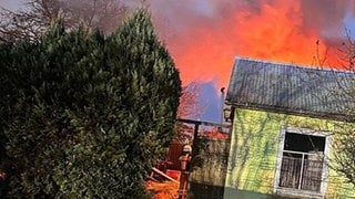 Hohe Flammen schlagen aus einer Kleingartenanlage am Flughafen Speyer. Feuerwehrmänner bekämpfen die Flammen.