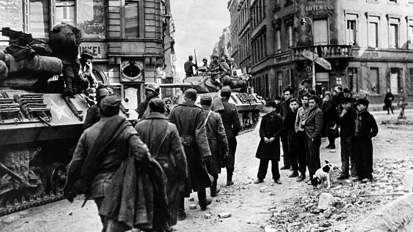 Eine Gruppe Jugendlicher steht am Straßenrand (r) und beobachtet den Einmarsch von US-Soldaten und Panzern in Mainz gegen Ende des Zweiten Weltkriegs.