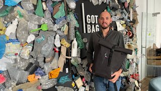 Got Bag-Gründer Benjamin Mandos steht mit einem Rucksack vor einer mit Plastik beklebten Wand