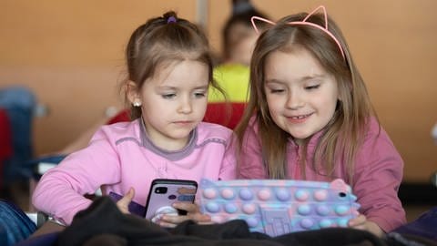 Zwei kleine Mädchen aus der Ukraine sitzen in der zu einer Notunterkunft umfunktionierten Turnhalle in Bad Kreuznach und spielen an einem Tablet.