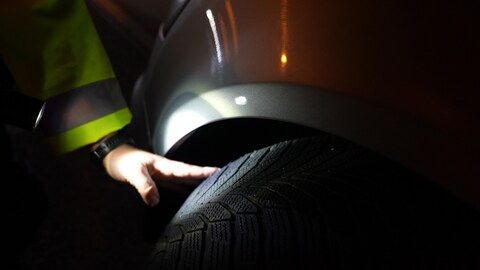 Die Hand eines Polizisten berührt einen Autoreifen und leuchtet mit einer Taschenlampe auf das Profil des Reifens.