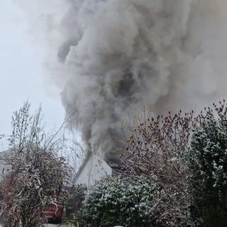 Schwere graue Rauchwolken steigen aus dem Dach eines brennnenden Hauses in Erbes-Büdesheim auf. Die Feuerwehr ist vor Ort und löscht. 