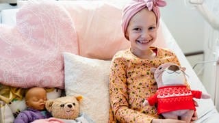 Emilia sitzt lächelns auf einem Krankenbett, in dem rosafarbene Kissen. Sie hält ein Plüschtier in den Händen und hat ein über der Stirn geknotetes Kopftuch auf. 