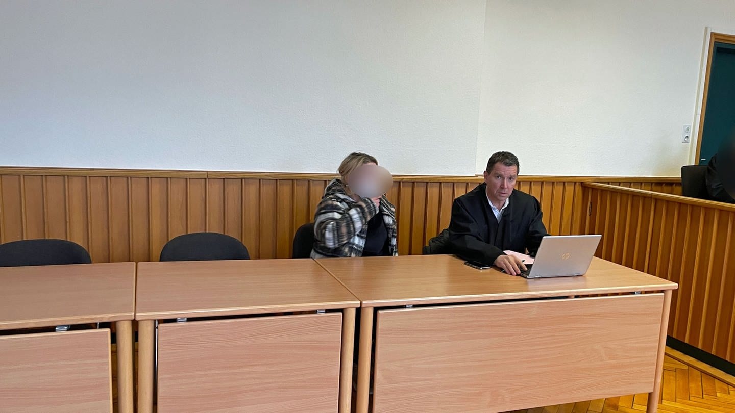 Eine Angeklagte sitzt mit Maske im Amtsgericht in Bad Sobernheim neben ihrem Anwalt. Sie hat blonde Strähnchen und längere Haare und trägt ein schwarzes T-Shirt und eine karierte Hemd-Jacke.