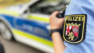 Eine Polizistin steht neben einem Streifenwagen. Auf ihrer Dienstkleidung ist das Wappen des Landes Rheinland-Pfalz zu sehen.