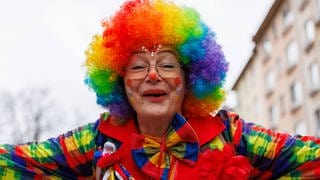 Eine Frau, die als bunter Clown verkleidet ist, breitet fröhlich ihre Arme aus. Sie ist beim Rosenmontagszug in Mainz 2024 mitgelaufen und gehört zum Stammtisch "Die Allerscheenste", das ist ein Verein aus dem rheinhessischen Lörzweiler. 