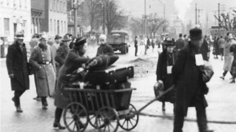 Ein Mann mit Schild um den Hals zieht eine Karre, die mit Koffern beladen ist, eine Frau hält die Koffer fest.