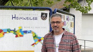 Ortsbürgermeister Setz steht vor einem Schild mit dem Logo von Heimweiler