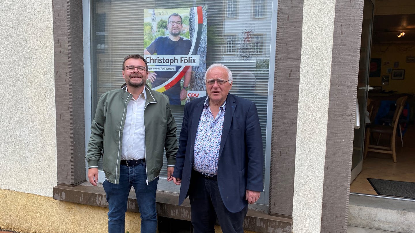 Christoph und Martin Fölix stehen vor einem Wahlplakat, das an einer Scheibe klebt.