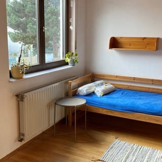 Blick in ein Zimmer mit Bett, Nachttisch und einem kleinen Regal an der Wand.
