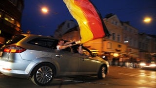 Feiernde Fans schwenken aus einem Auto heraus eine große Deutschland-Fahne.