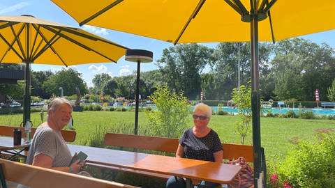 Zwei ältere Damen sitzen unter einem Sonnenschirm auf einer Biergarnitur und unterhalten sich - im Hintergrund ist das Schwimmerbecken des Freibades Gimbsheim.