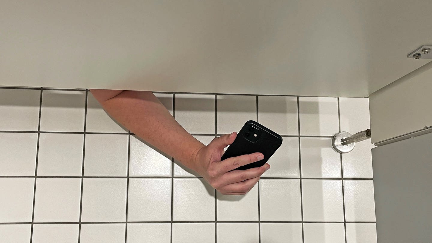 Ein Mann filmt mit seinem Handy unter der Toilettenkabine hindurch eine Frau.