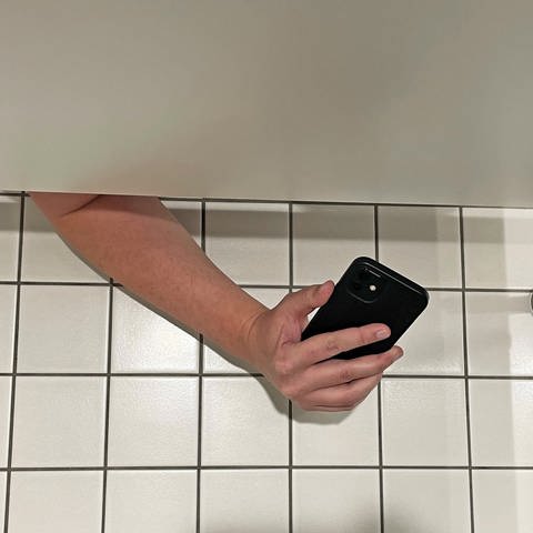 Ein Mann filmt mit seinem Handy unter der Toilettenkabine hindurch eine Frau.