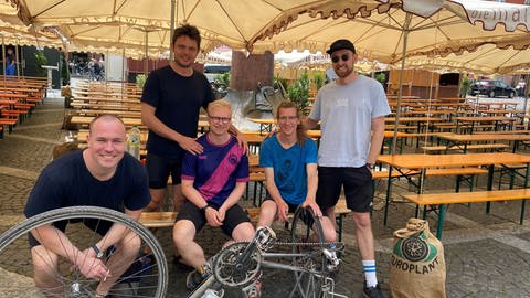 Vier Männer unter Sonnenschirmen stehen und sitzen hinter einem Fahrrad, das umgedreht auf dem Sattel steht.