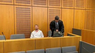 Der Angeklagte sitzt auf der Anklagebank, neben ihm steht sein Verteidiger.