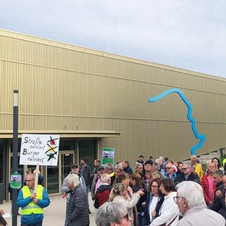 Vor der Rheinhessenhalle in Monsheim stehen Menschen und halten Protest-Schilder hoch.