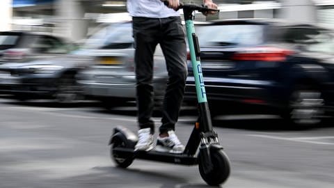 Ein E-Scooter Fahrer rast mit hoher Geschwindigkeit auf der Straße an parkenden Autos vorbei. 