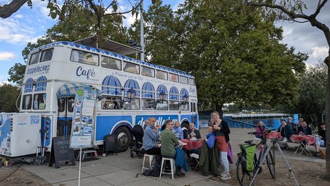 Helene, eine Frau mit blondem Haar steht vor dem großen Bus und trägt Kaffee an eine Gruppe von Menschen aus. 