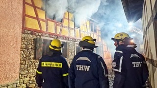 Helfer des THW stehen vor dem Fachwerkhaus in Wörrstadt. Aus den Fenstern steigt dichter Rauch auf.