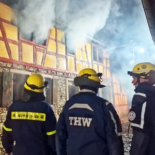 Helfer des THW stehen vor dem Fachwerkhaus in Wörrstadt. Aus den Fenstern steigt dichter Rauch auf.