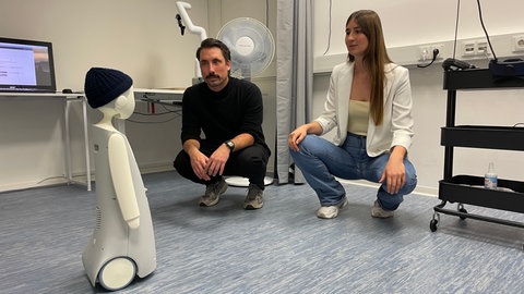 Ein kleiner Roboter steht vor dem Boden. Vor ihm kniet ein Mann und eine Frau. Sie sprechen mit ihm.