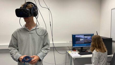 Links im Bild steht ein junger Mann, der eine VR-Brille trägt. Rechts im Bild sitzt eine junge Frau auf dem Computer und steuert das Programm, dass den Roboter in der VR-Simulation fahren lässt. 