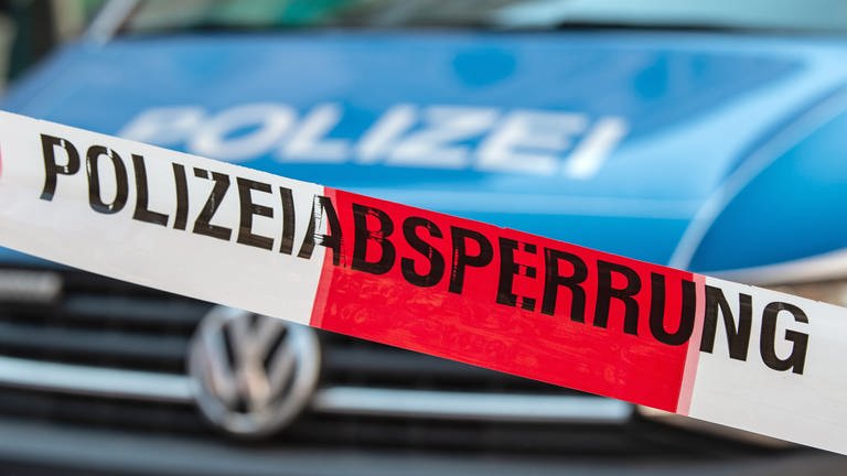Ein Band mit der Aufschrift "Polizeiabsperrung" ist vor einem Polizeiauto gespannt. Die Kriminalpolizei ermittelt aktuell nach dem Fund von zwei Leichen in Bitburg. Bei den Toten handelt es sich wohl um ein geschiedenes Ehepaar.