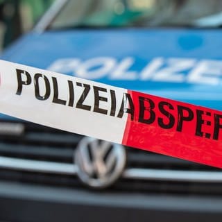 Ein Band mit der Aufschrift "Polizeiabsperrung" ist vor einem Polizeiauto gespannt. Die Kriminalpolizei ermittelt aktuell nach dem Fund von zwei Leichen in Bitburg. Bei den Toten handelt es sich wohl um ein geschiedenes Ehepaar.
