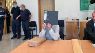 Der Angeklagter sitzt im Gerichtssaal beim Prozess in Idar-Oberstein. Er verdeckt mit einem Aktenordner sein Gesicht. Dem 55-Jährigen wird vorgeworfen, im Februar 2022 im Internet dazu aufgerufen zu haben, Polizisten zu erschießen.