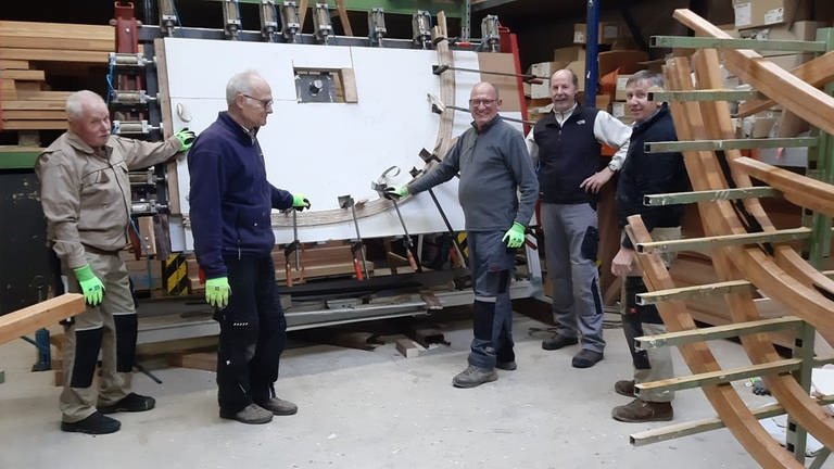 Stolz präsentieren die Mitglieder des Fördervereins Neumagener Weinschiff ihre Arbeit. Mit Hydraulikdruck werden die Leisten zur Herstellung der neuen Querverbindungen für das Schiff vorgebogen. Sie bleiben , bis der Leim abgebunden hat, etwa sechs Stunden in der pneumatischen Presse. Danach behalten sie die gebogene Form bei. 