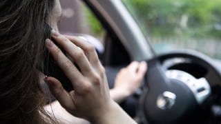Eine Frau sitzt mit Handy am Steuer. Das Amtsgericht Trier hat Einsprüche von drei Autofahrern gegen Bußgeldbescheide wegen Nutzung eines Mobiltelefons