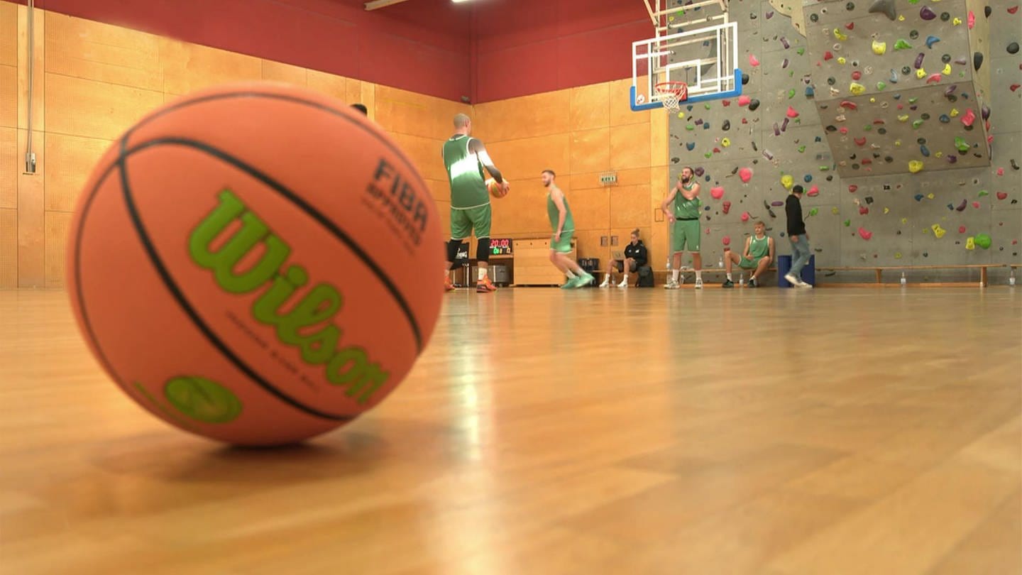 Basketball liegt auf dem Boden einer Sporthalle, im Hintergrund Basketballspieler und ein Korb