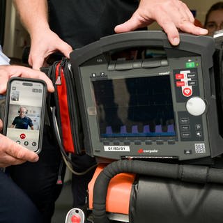 Neben ein mobiles EKG-Gerät wird ein Handy gehalten. Auf diesem läuft eine Videoschalte mit einem Telenotarzt. In Rheinland-Pfalz wird nun ein zweiter Telenotarzt-Standort eröffnet, nämlich in Trier.