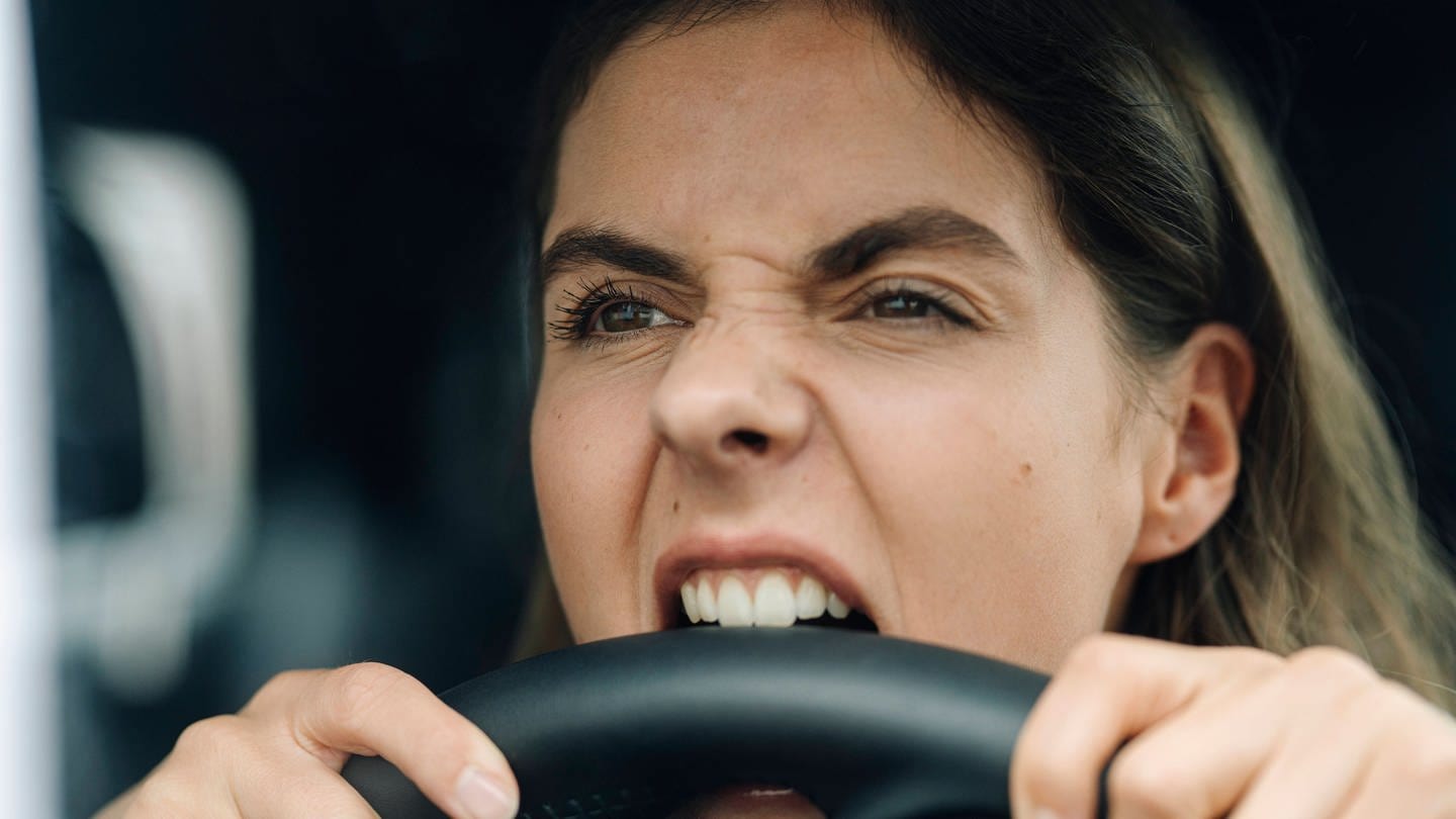 Nahaufnahme einer wütenden Frau, die ins Lenkrad beißt, während sie in einem Auto sitzt