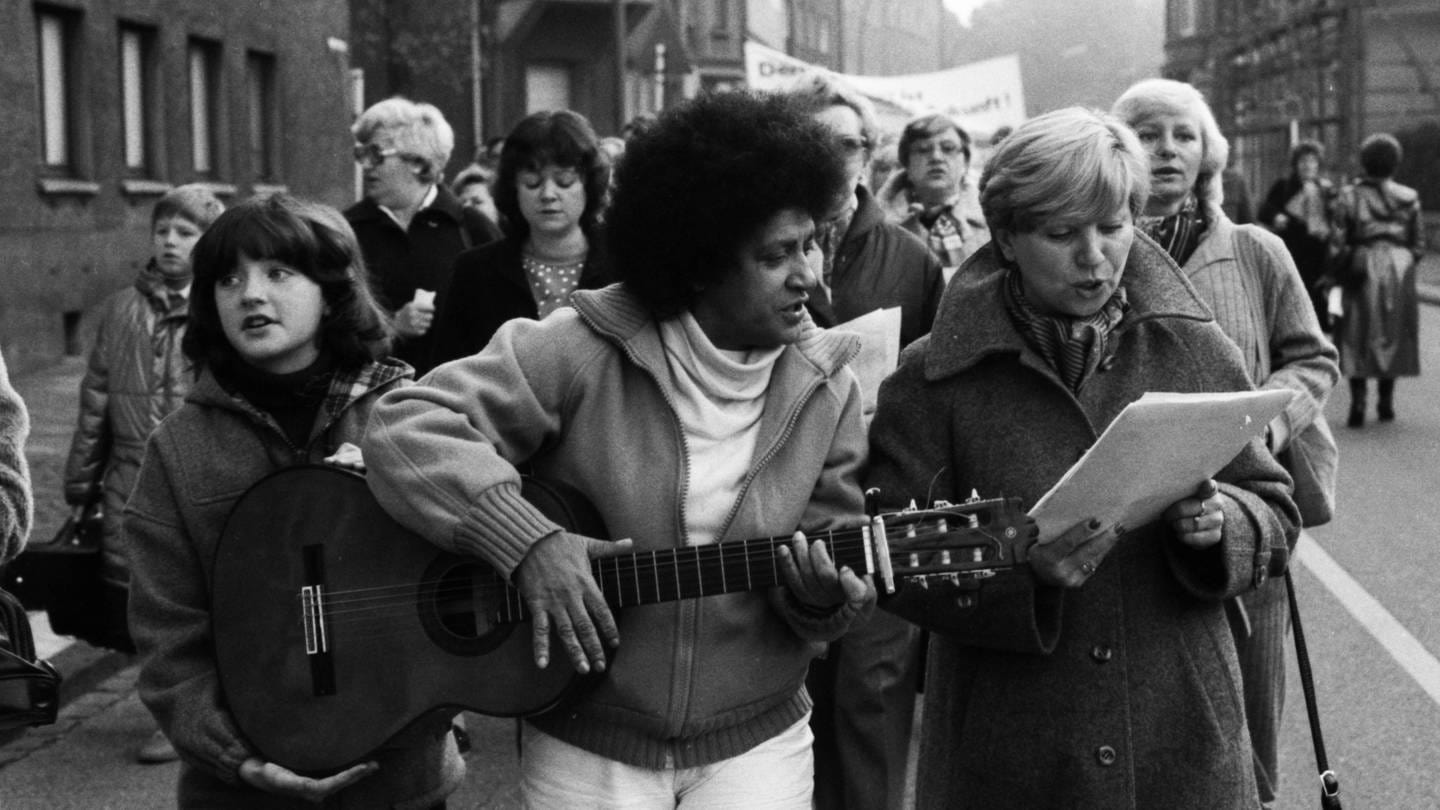 Fasia Jansen bei einer Demo mit Gitarre. Sie schaut auf ein Blatt, das ihr eine Frau im Mantel zeigt.