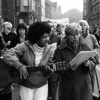 Fasia Jansen bei einer Demo mit Gitarre. Sie schaut auf ein Blatt, das ihr eine Frau im Mantel zeigt.