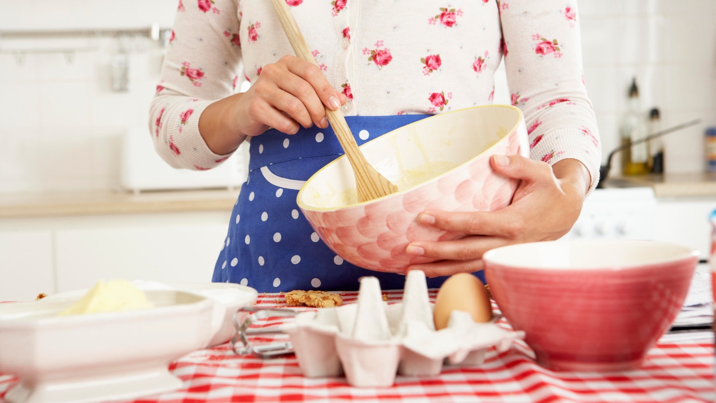 Junge Frau in Blümchenbluse und blauer Schürze rührt mit einem Holzlöffel in einer rosa Schüssel einen Kuchenteig an: #Tradwife, Abkürzung für 