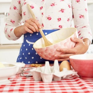 Junge Frau in Blümchenbluse und blauer Schürze rührt mit einem Holzlöffel in einer rosa Schüssel einen Kuchenteig an: #Tradwife, Abkürzung für "traditionelle Ehefrauen", und #Stayathomegirlfriend haben in den vergangenen Jahren TikTok und Instagram erobert. Die Message: Nichts ist erfüllender für eine Frau, als für einen Mann zu sorgen und die Kinder großzuziehen. 