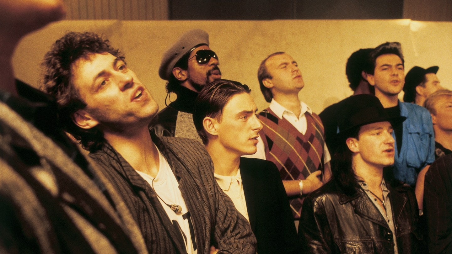 Sir Bob Geldof (links), Paul Weller, Robert Kool Bell, Phil Collins und Bono sowie andere Künstler stehen nebeneinander und singen, 1984.