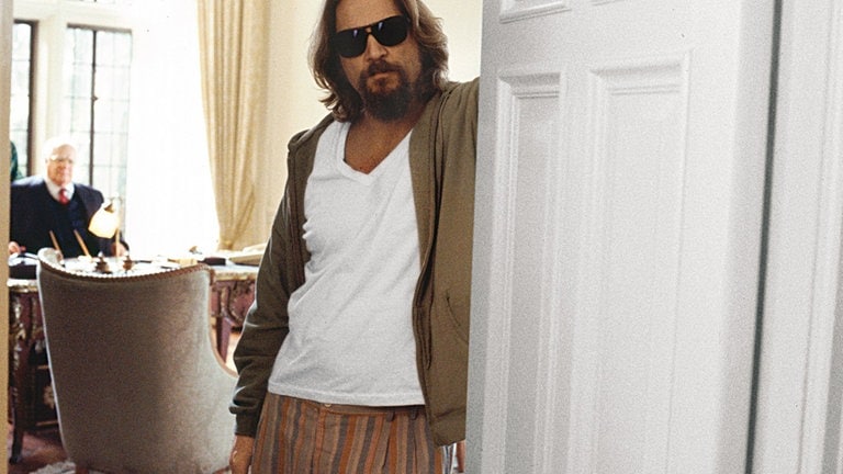 Jeff Bridges in seiner Rolle als Jeffrey Lebwoski steht in einem Türrahmen. Er trägt ein weißes T-Shirt, Strickjacke und Sonnenbrille. 