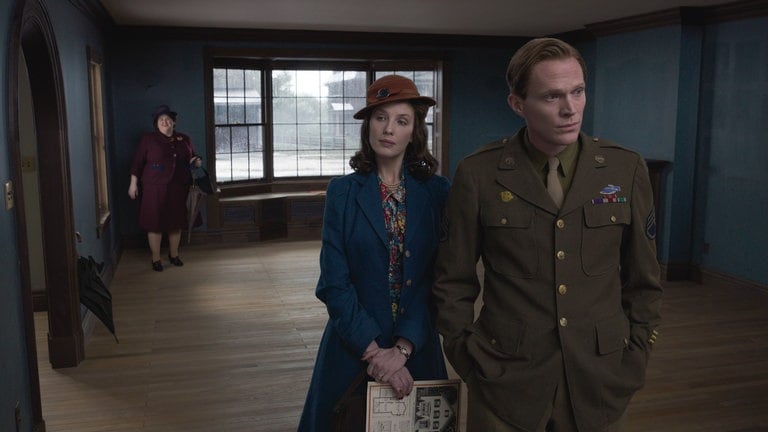 Das Ehepaar Young (Kelly Reilly, Mitte, und Paul Bettany, rechts) steht im Film „Here“ in einem großen Wohnzimmer. Er trägt eine Militäruniform. Im Hintergrund ist eine Maklerin zu sehen.