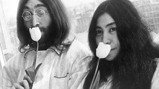 Auf einem schwarz-weißen Foto sind ein junger John Lennon (links) und Yoko Ono (rechts) zu sehen. Sie riechen an einer Tulpe.