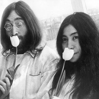 Auf einem schwarz-weißen Foto sind ein junger John Lennon (links) und Yoko Ono (rechts) zu sehen. Sie riechen an einer Tulpe.