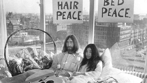 John Lennon (links) und Yoko Ono sitzen in einem Bett, im Hintergrund ist Amsterdam zu sehen. 