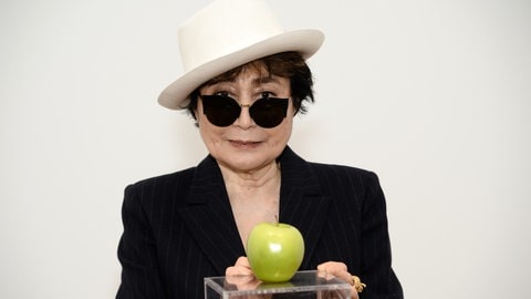 Die Künsterlin Yoko Ono trägt einen Hut und Sonnenbrille und zeigt ihr Kunstwerk „Apple“ von 1966.